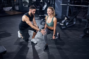 Personal trainer wearing dark clothes fixing the leg position of a female athlete before the start of dumbbell activity.